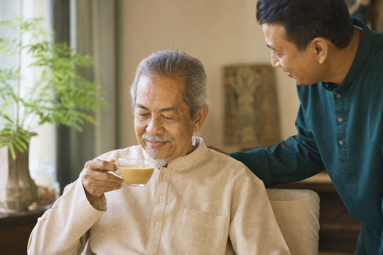 An elderly Malaysian parent enjoying a warm golden drink in soft light with an adult child nearby, dignified caring warm mood